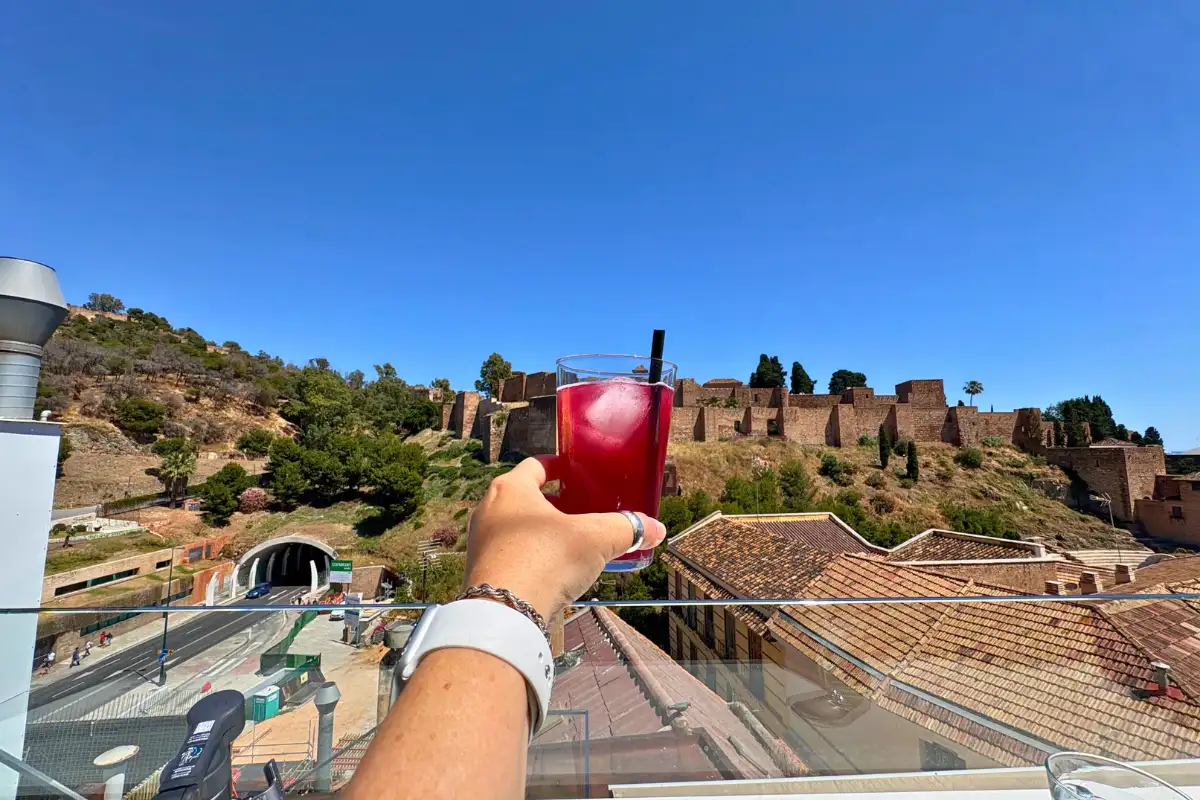 Hand holding a red cocktail over rooftops toward the Alcazaba in Malaga